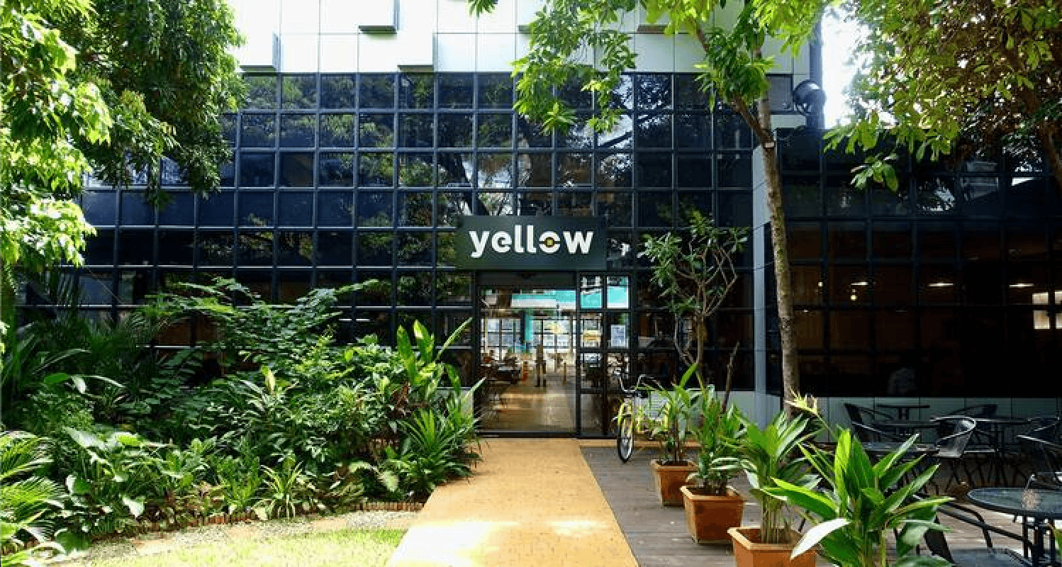 Glass-front entrance to Yellow coworking space with lush garden walkway and ‘yellow’ sign.