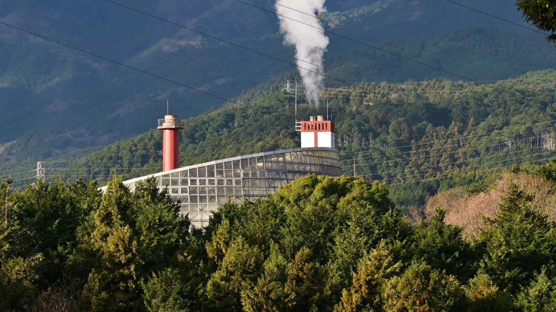 Industrial facility with two smokestacks, one emitting white steam, set against forested hills and mountains.