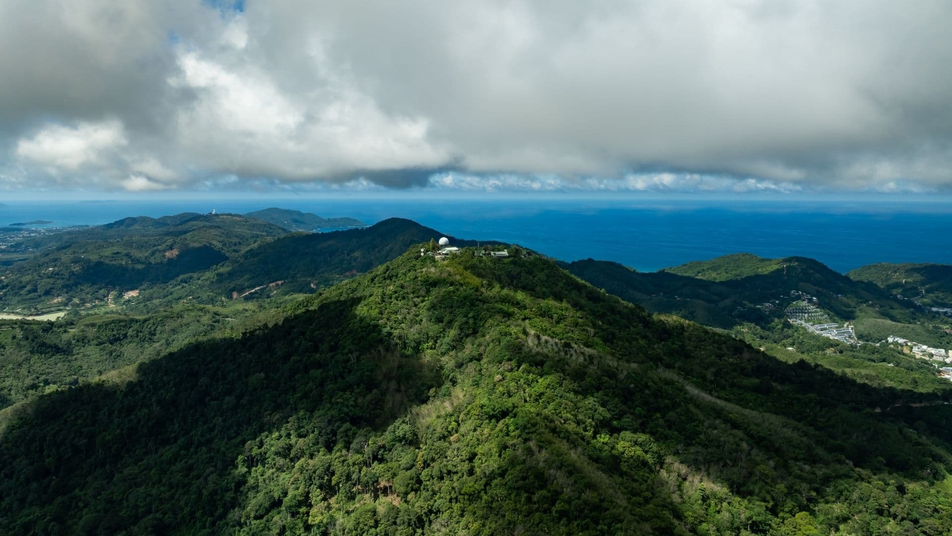 Aerial view of dense green forested mountains meeting a deep blue sea, with a white radar dome station on the central peak and a heavy grey cloud bank across the horizon.