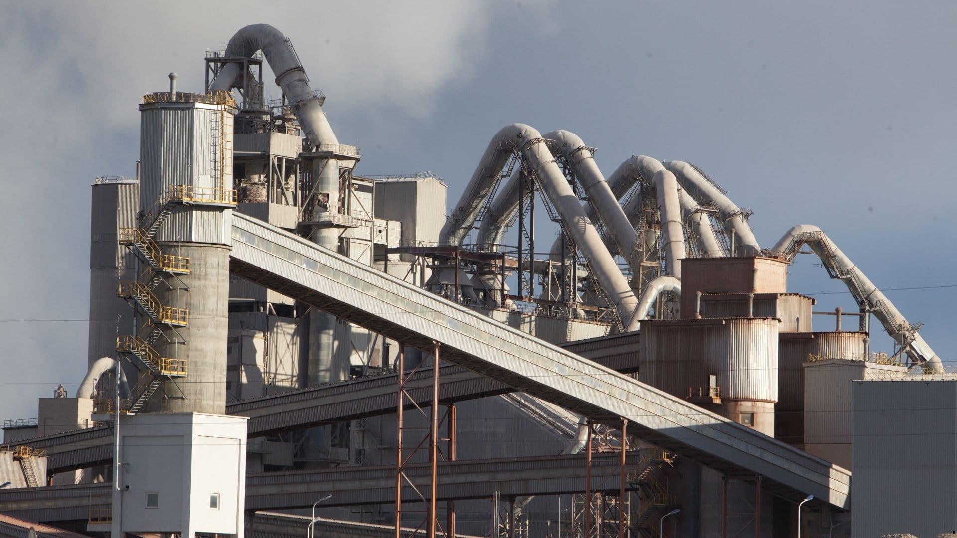 Cement plant preheater tower with cyclone ducts and conveyor galleries under a cloudy sky